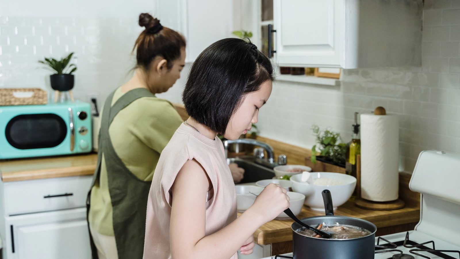 A young person stirring a pot on the stove in a home kitchen while an adult works at the sink behind them.