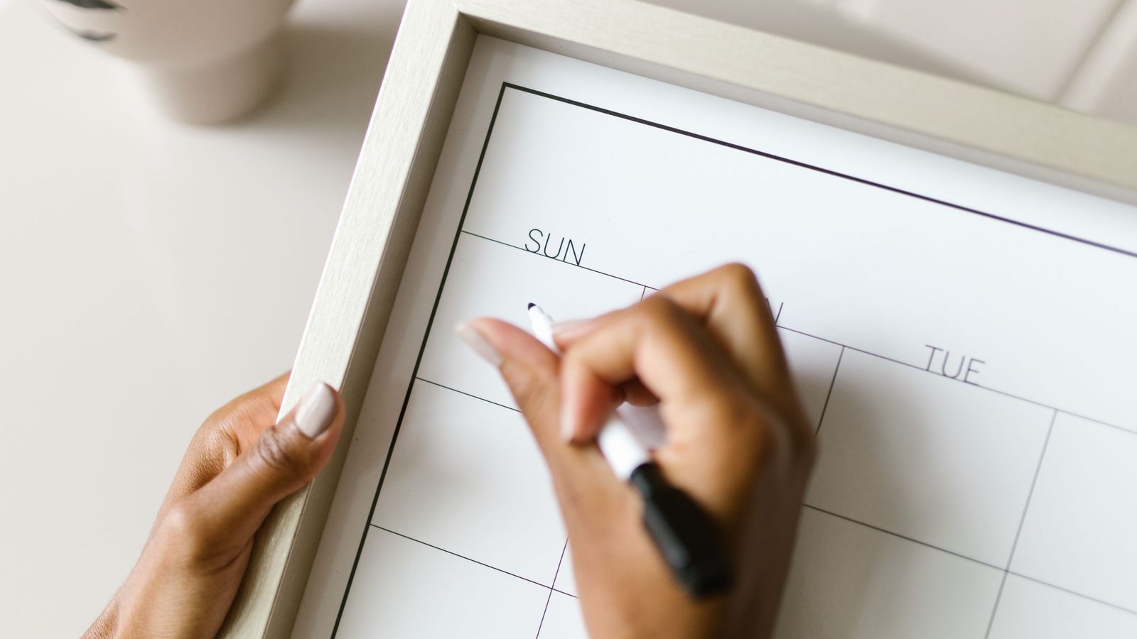 Hands write on a dry-erase weekly calendar, starting at the Sunday square.