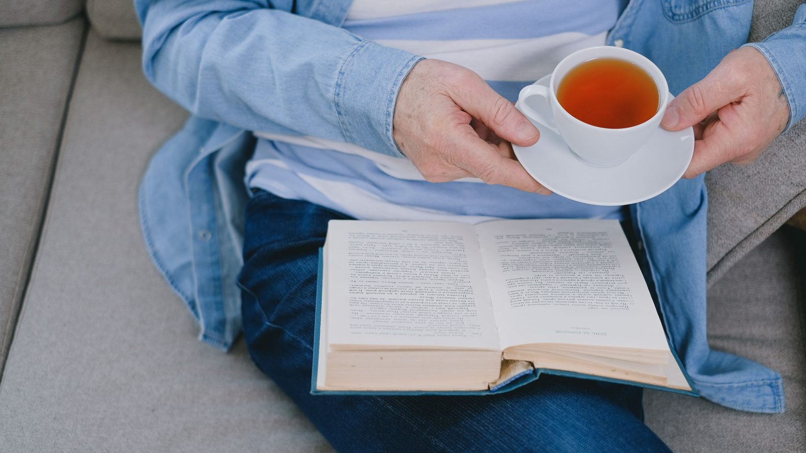 A person on a sofa holds a cup of tea over an open book in their lap.