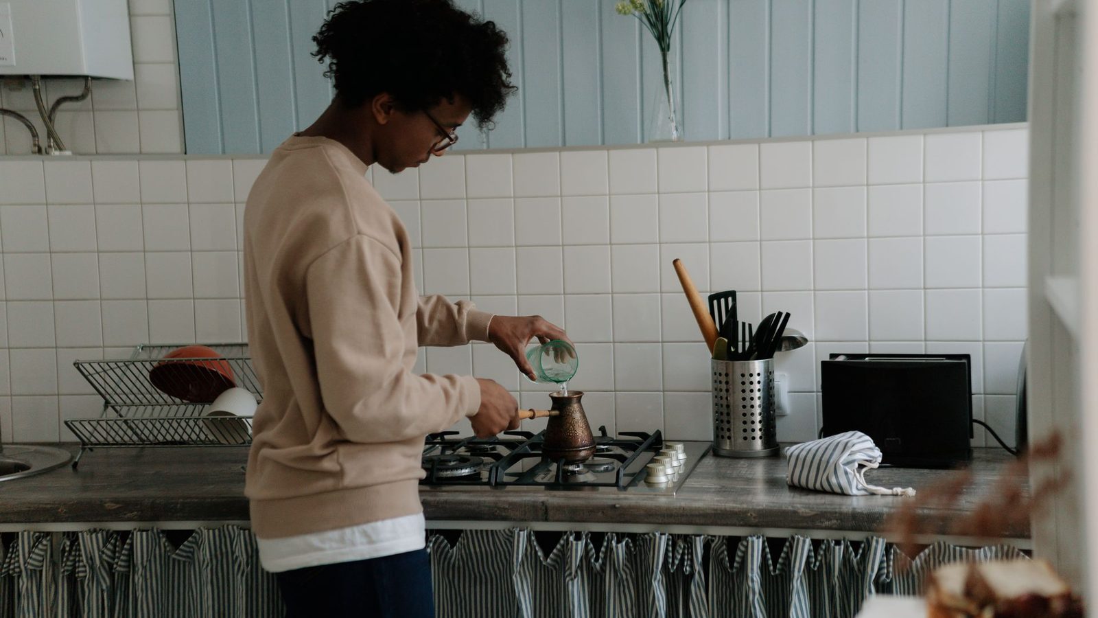 A young person in glasses pours water into a small coffee pot on a gas stove in a home kitchen.